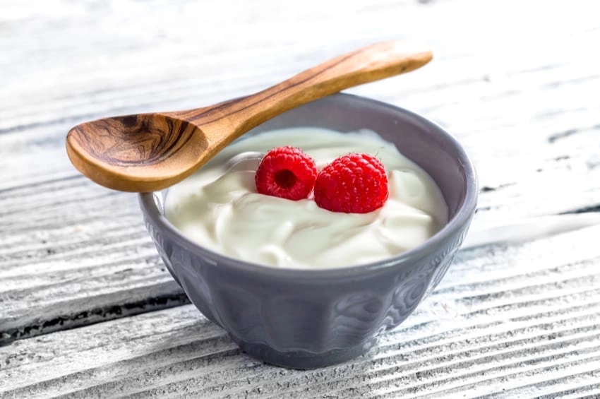 Bowl of Greek yogurt topped with two raspberries and a wooden spoon resting on the rim on a rustic wooden surface.